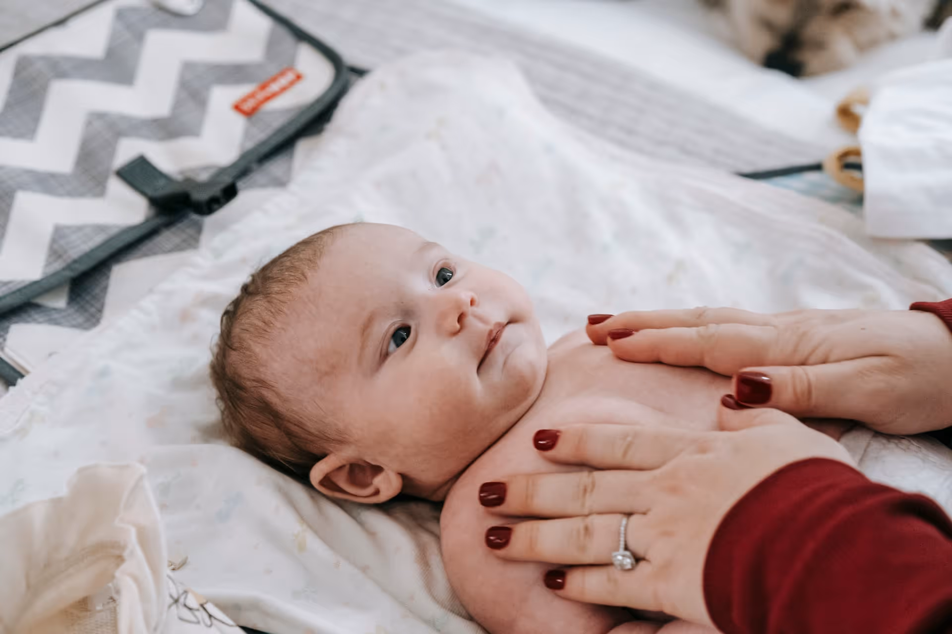 A baby on a changing mat, a parent&rsquo;s hand resting on their chest.